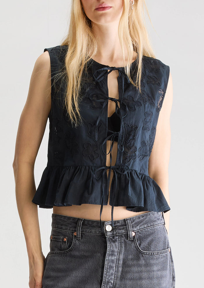 A woman wearing a sleeveless tie front top in black with tonal lacework detailing and peplum hem.