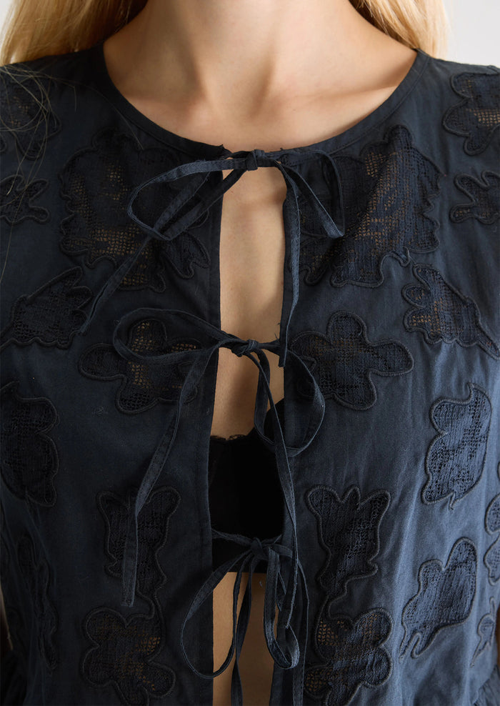 A woman wearing a sleeveless tie front top in black with tonal lacework detailing and peplum hem.