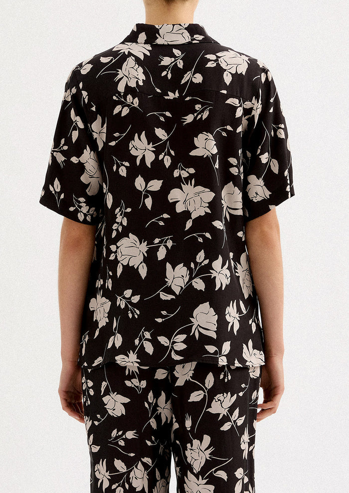 A person wearing a short sleeve collared shirt in black with white floral pattern.