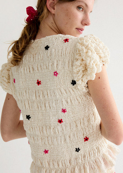 A woman wearing a smocked and ruffled cream top with red and pink embroidered flowers throughout.