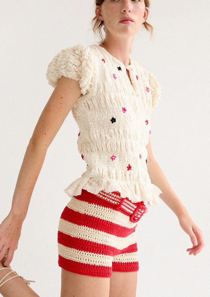 A woman wearing a smocked and ruffled cream top with red and pink embroidered flowers throughout.