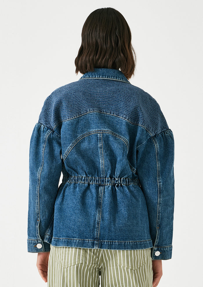 A woman wearing a denim jacket with decorative stitching and ruched shoulder detail.