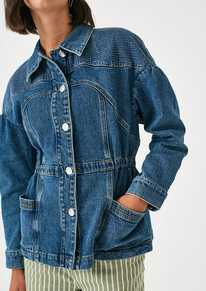 A woman wearing a denim jacket with decorative stitching and ruched shoulder detail.
