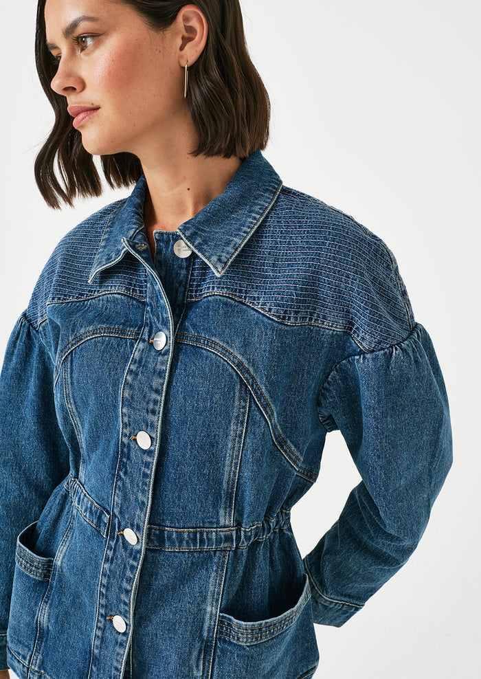 A woman wearing a denim jacket with decorative stitching and ruched shoulder detail.