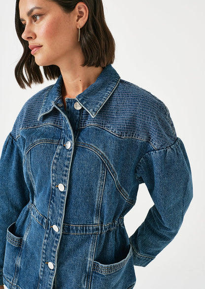 A woman wearing a denim jacket with decorative stitching and ruched shoulder detail.